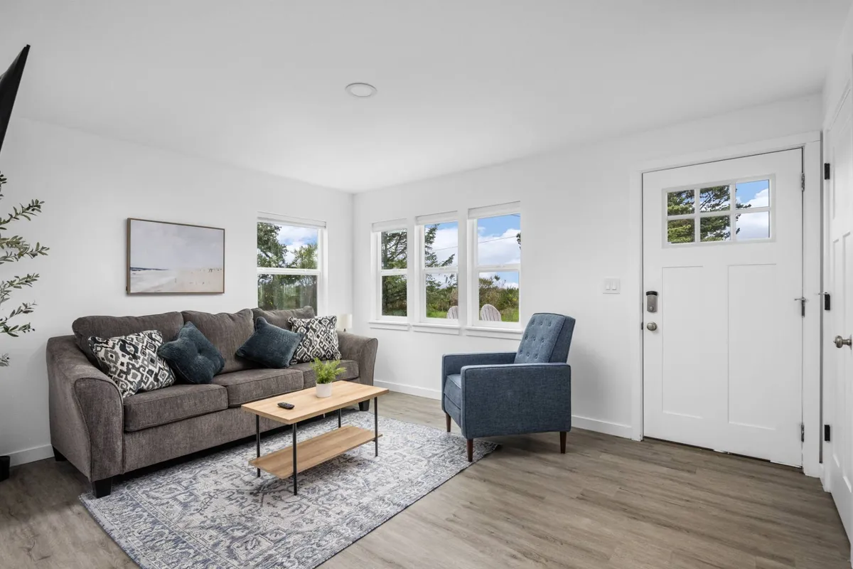 The image showcases a cozy living area with a comfortable sofa and armchair arranged around a coffee table, inviting guests to relax and unwind. Large windows frame the space, filling it with natural light and offering glimpses of the scenic coastal views. Thoughtful décor and artwork add warmth and character, while the layout encourages socializing, reading, or simply enjoying the serene ambiance.