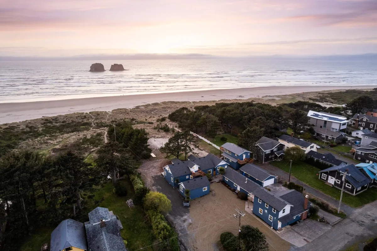 A wide-angle view of Rockaway Beach with the ocean stretching out, sandy shoreline, distant rock formations (likely Twin Rocks Arch), and nearby houses. The setting appears serene, with soft lighting suggesting either early morning or sunset. The natural landscape is prominent and inviting for walks, exploration, and relaxation.