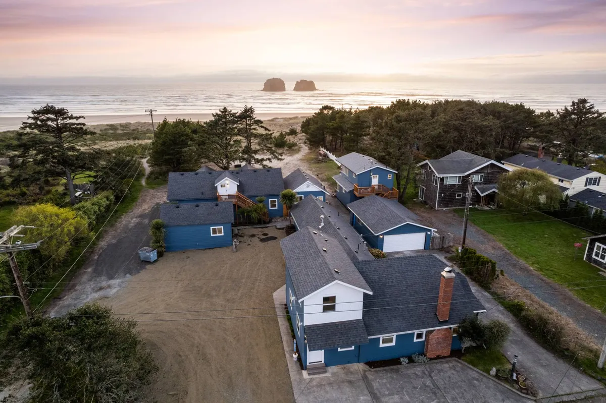 An exterior scene showing a group of houses nestled near the beach, with a view of the ocean and vivid sunset on the horizon. People are seen gathering, walking, and enjoying the outdoors, evoking a relaxed and communal atmosphere. Large trees frame the scene, adding natural beauty.