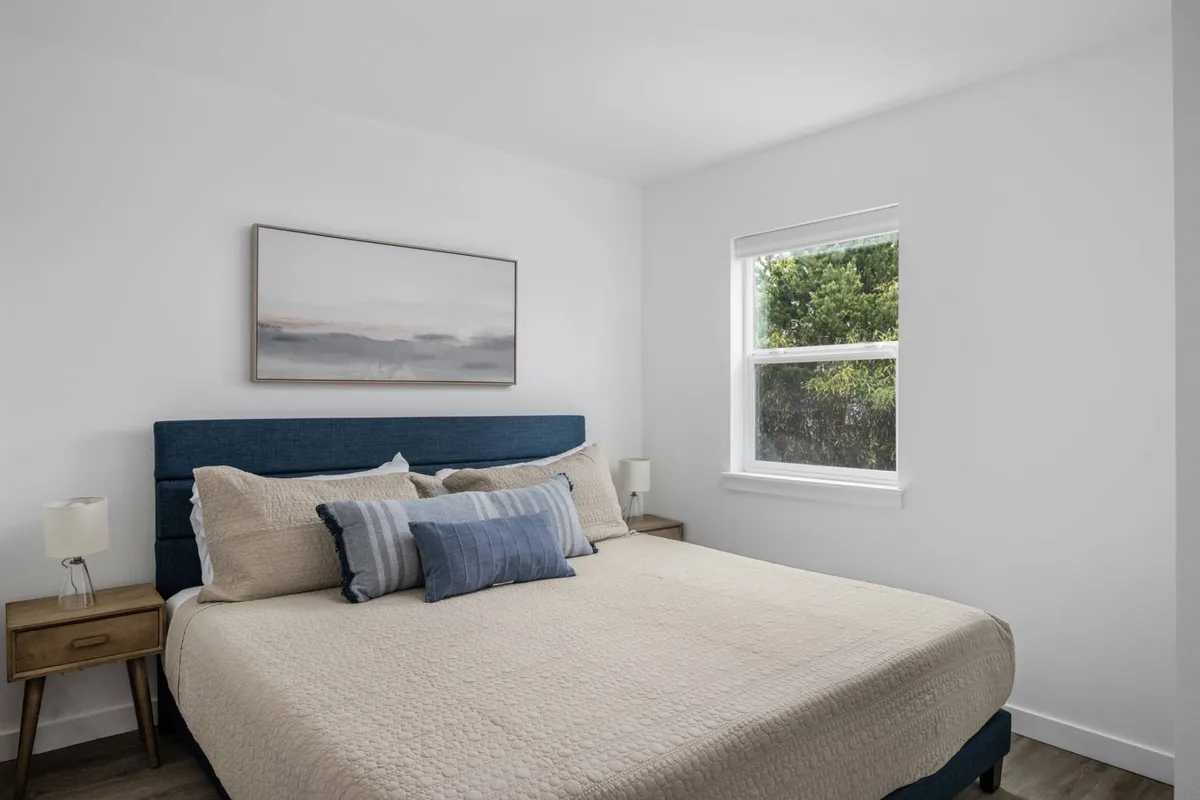 A cozy bedroom interior with a made bed, large window providing natural light, tasteful artwork, and nightstands on either side. The space feels peaceful and perfect for relaxation or a slow morning.