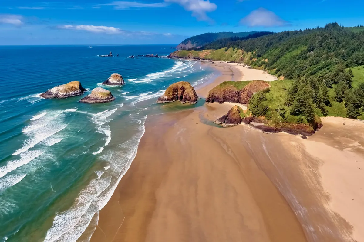 This image captures an expansive sandy beach bordered by iconic rock formations, with gentle ocean waves rolling in and distant hills under a tranquil sky. The scene is quiet and inviting, suggesting the restful, inspiring natural environment that makes working remotely at Ocean Rogue Inn so appealing.
