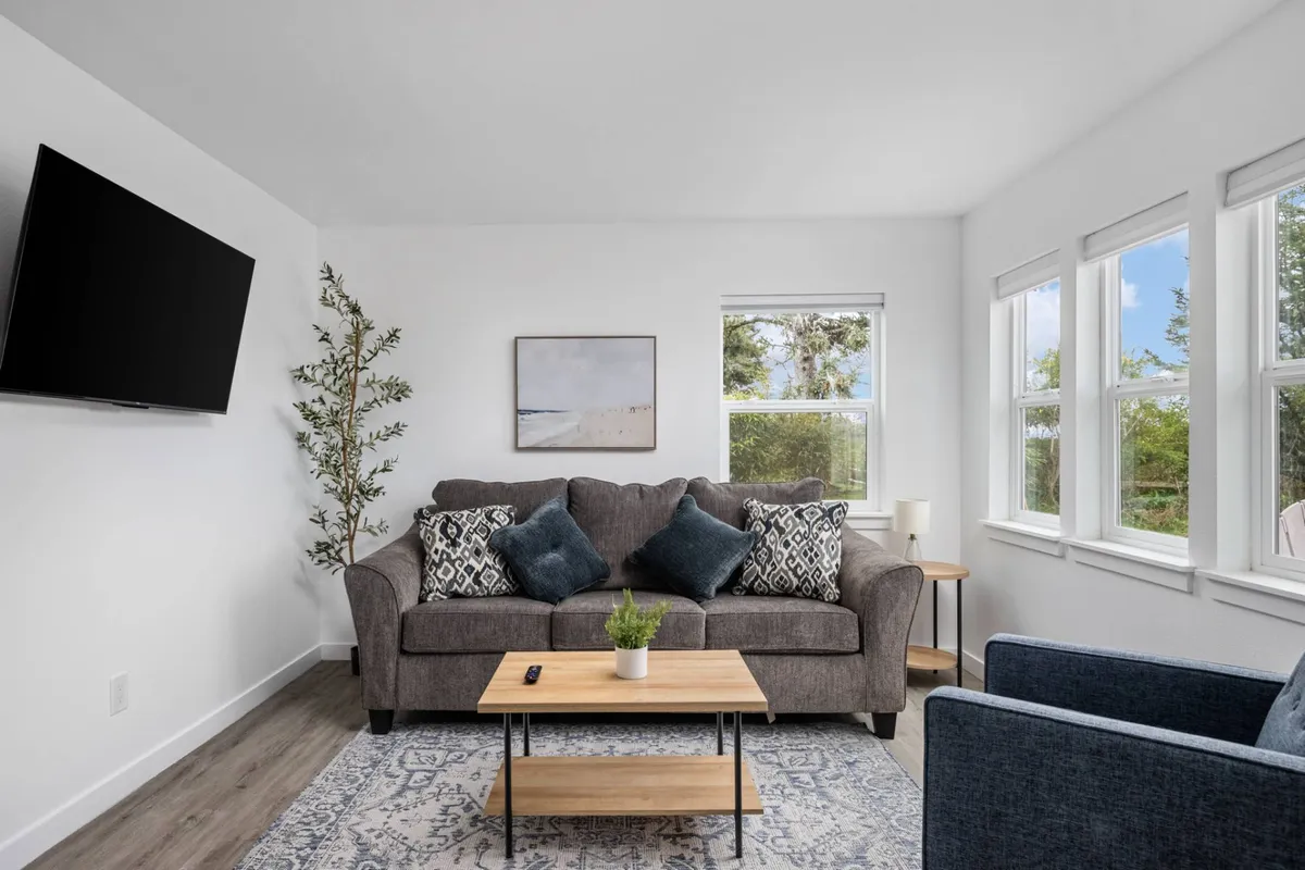 A cozy living room featuring a plush sofa and coffee table, positioned near large windows that let in ample natural light. Modern artwork decorates the walls, and a TV is set up for entertainment. The overall atmosphere is warm and inviting, perfect for relaxing or socializing.
